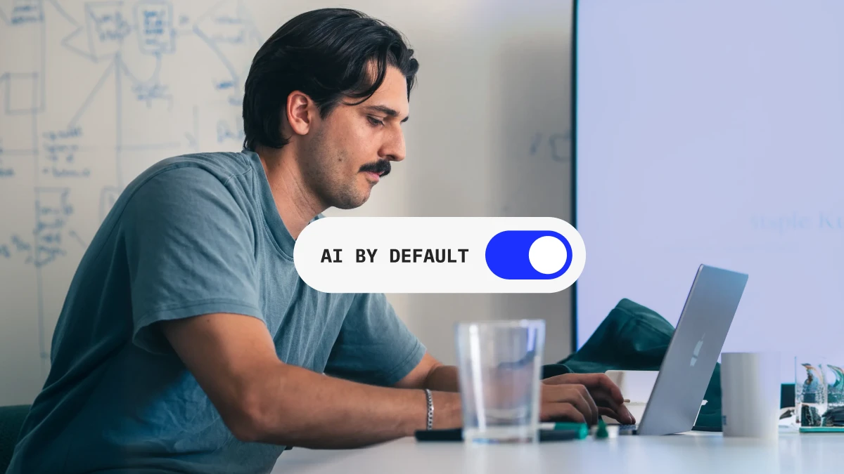 Ein Mitarbeiter arbeitet konzentriert am Laptop vor einem beschriebenen Whiteboard, davor ein eingeschalteter Schalter mit der Aufschrift „AI by Default“