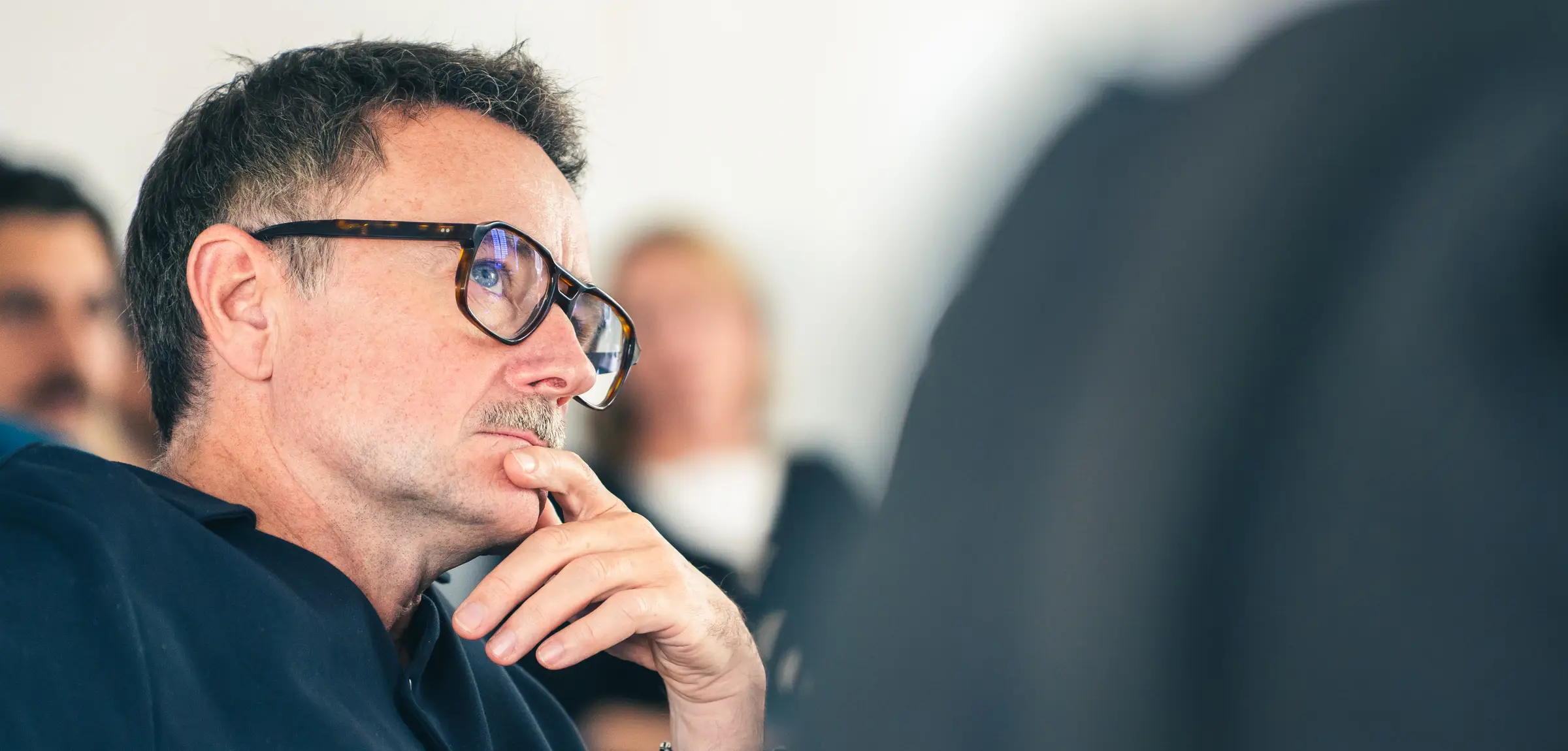 Ein grauhaariger Mann mit Brille stützt nachdenklich die Hand ans Kinn, hinter ihm unscharf weitere Menschen.